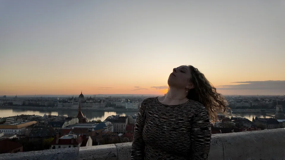 A person tipping their head back beside a city overlook at sunset