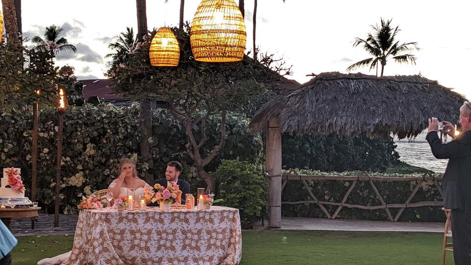 Two people seated at a flower-covered table under hanging woven lamps